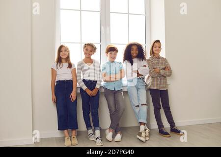 Gruppo di bambini felici e diversi in piedi vicino alla finestra, guardando la macchina fotografica e sorridendo Foto Stock