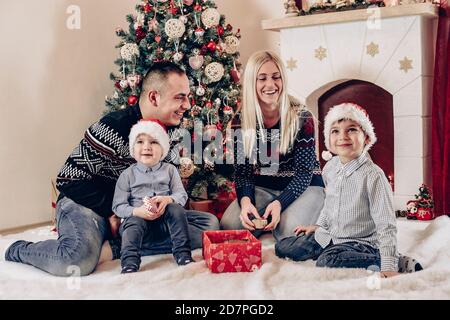 Madre, padre e bambini festeggiano seduti davanti all'albero di Natale, giocando e aprendo i regali - buona famiglia festeggia il Natale Foto Stock