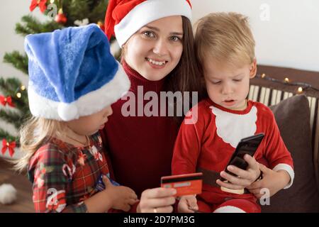 Natale, inverno, anno nuovo, Celebration, concetto di famiglia - mamma gioiosa fare shopping online con due piccoli bambini divertenti sedersi vicino albero di Natale tenere sconto Foto Stock