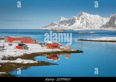 Casale rosso riflesso nel fiordo Rossoy Straumen, nel lontano oceano posteriore, montagne innevate Hustinden e Bjoerntinden, Nordland, Lofoten, Norvegia Foto Stock