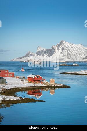 Casale rosso riflesso nel fiordo Rossoy Straumen, nel lontano oceano posteriore, montagne innevate Hustinden e Bjoerntinden, Nordland, Lofoten, Norvegia Foto Stock