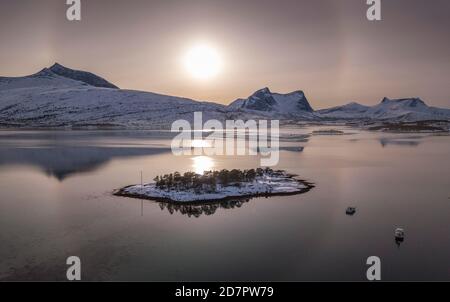 Anello del sole, alone su catena montuosa innevata, paesaggio artico invernale, di fronte piccola isola e barche da pesca in un fiordo, Ballangen, Nordland, Norvegia Foto Stock