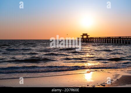 Napoli, Florida rosso giallo e arancio tramonto nel golfo della costa del Messico con il sentiero del sole dal molo in legno con orizzonte e blu scuro onde oceano Foto Stock