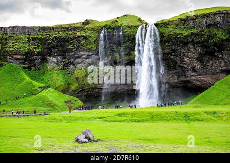 Seljalandsfoss, cascata islandese con scogliera d'acqua bianca in verde estate paesaggio roccioso e le persone a piedi sul sentiero Foto Stock