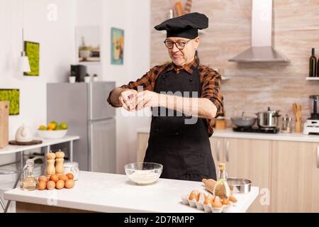 Uomo anziano che cricchiola le uova sulla farina di grano mentre prepara cibo delizioso. Anziano pasticciere spezzare l'uovo su una ciotola di vetro per la ricetta della torta in cucina, mescolando a mano, impastando gli ingredienti prreparing torta fatta in casa Foto Stock