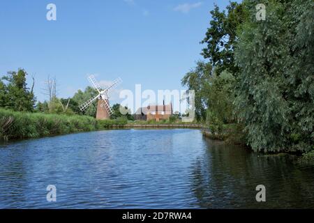 Un mulino a vento tradizionale (pompa del vento) accanto ad un cottage o una casa graziosa su una curva in un fiume nel Norfolk Broads, Inghilterra, Regno Unito. Giorno di sole, acqua blu AN Foto Stock