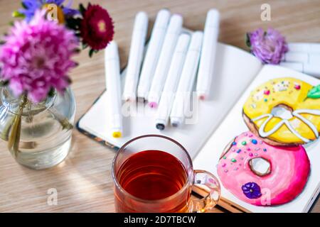 Tazza di tè trasparente su fondo ciambelle di schizzo. Il tutto posto su un tavolo di legno con una tazza trasparente di tè, taccuino, pennarelli e fiori. Hobby. Ciambelle da disegno. Messa a fuoco morbida Foto Stock