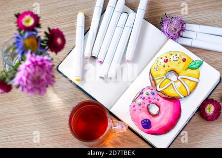 Disegno di ciambelle nel notebook. Il tutto posto su un tavolo di legno con una tazza trasparente di tè, taccuino, pennarelli e fiori. Hobby. Ciambelle da disegno. Vista dall'alto. Messa a fuoco morbida Foto Stock