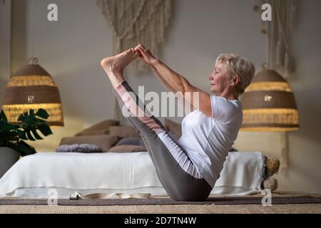 Donna anziana attiva che fa l'addestramento di yoga a casa, tenendo la navasana o la posa di yoga della barca Foto Stock