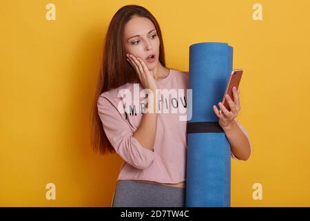 Studio di sorprendente ragazza dai capelli scuri con corpo perfetto, tenendo in mano yoga tappetino e moderno smartphone, lettura messaggio scioccato, tenendo la bocca Foto Stock