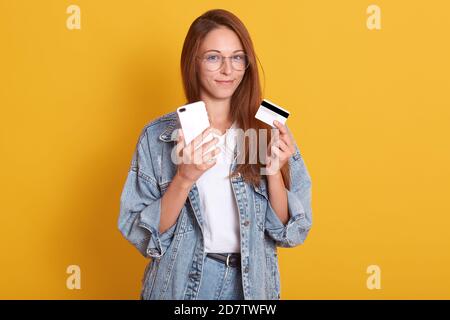 Tiro orizzontale di adorabile donna sorridente wering abito casual isolato su sfondo giallo, tenendo gadget e carta di credito in mani, facendo online Foto Stock