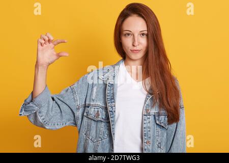 Studio shot di bella donna dai capelli scuri indossa una giacca demin mostra qualcosa di piccolo con la mano, isolato su sfondo giallo. Bello da giovane Foto Stock