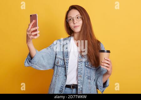 Ritratto di piacevole tenero bella fare selfie, tenendo smartphone e tazza di carta di caffè, indossando t-shirt bianca e giacca jeans, pranzo, it Foto Stock