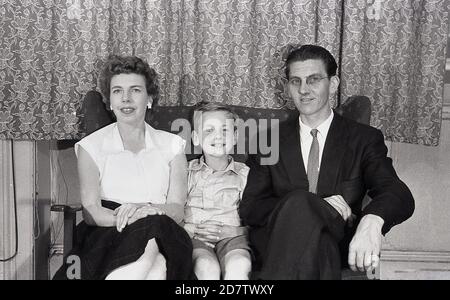 1950s, immagine storica di famiglia di madre, padre e figlio seduti insieme su una poltrona. Entrambi gli adulti sono vestiti formalmente con la signora che indossa una gonna di controllo e l'uomo che indossa un vestito & cravatta, come sarebbe stato il caso in questa era, anche a casa. Foto Stock