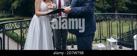 Coppia nuziale firma ufficiale certificato di matrimonio nelle mani di un registrar. La sposa giovane indossa un abito bianco e tiene un bouquet di nozze con rose rosa. CLA Foto Stock