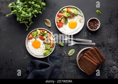 Uova fritte con insalata di micro germogli verdi di avocado di pomodoro per colazione su fondo nero di cemento, vista dall'alto Foto Stock