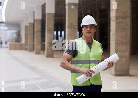 Ritratto in vita di un lavoratore di costruzione professionale che tiene piani e guardando la fotocamera mentre si trova in piedi in ufficio edificio, copia spazio Foto Stock