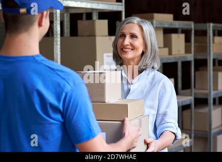 Felice manager donna matura che dà pacchi scatole al corriere in magazzino. Foto Stock