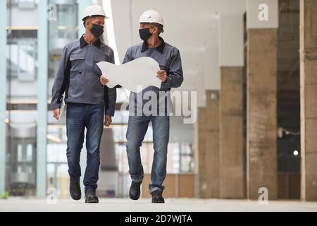 Ritratto a lunghezza intera di due lavoratori che indossano maschere e che tengono piani mentre si cammina verso la fotocamera in cantiere, copia spazio Foto Stock