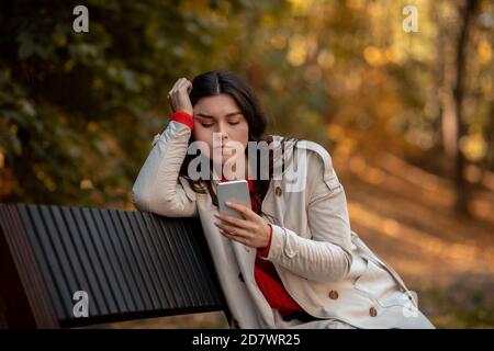Triste giovane donna in autunno vestito seduta su panchina a. parcheggiare con lo smartphone e sentirsi depresso Foto Stock