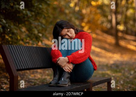 Giovane donna depressa che le aggira le ginocchia mentre si siede sulla panchina al parco giallo d'autunno Foto Stock