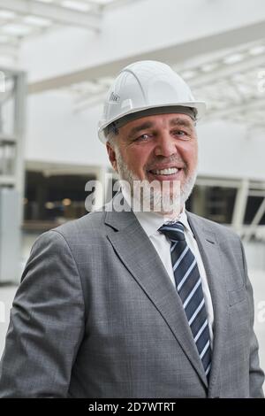 Ritratto verticale di un uomo d'affari sorridente che indossa un cappello e guarda in camera mentre si è in piedi in cantiere Foto Stock