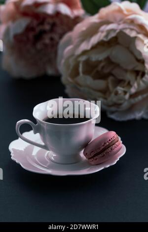 Caffè mattutino in una tazza di porcellana con macaron rosa e fiori di peon pastello. Messa a fuoco selettiva. Modalità oscura. Foto Stock