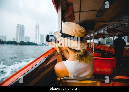 Giovane donna viaggiatore che scatta foto con il suo viaggio mobile a. Thailandia con cappello sulla barca a Bangkok Thailandia Foto Stock