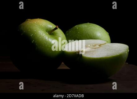 Still life, mela verde a metà e mele verdi con gocce d'acqua sulla cima in un chiaroscuro, stile barocco con sfondo nero su un tavolo in legno d'epoca Foto Stock