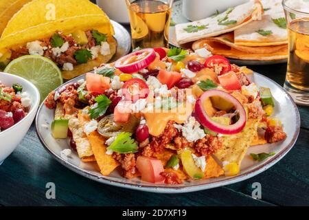 Cibo messicano. Nachos con manzo, fagioli, peperoncini, cipolle e salsa al formaggio da vicino, con tacos di maiale, quesadilla e tequila sullo sfondo Foto Stock