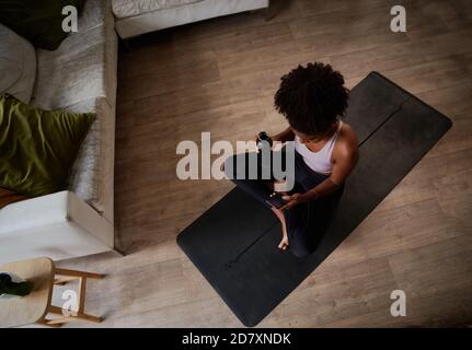 Vista ad alto angolo della giovane donna africana che tiene la bottiglia e. uso del telefono seduto sul tappetino yoga Foto Stock
