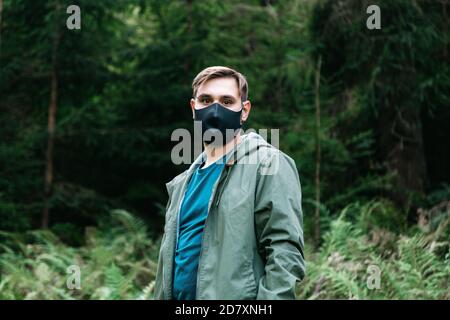 Uomo in maschera medica protettiva nera fuori in foresta da solo. L'uomo viaggia da solo mantenendo la distanza sociale nel turismo locale. Protezione contro Foto Stock