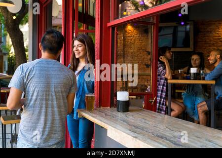 Un bell'uomo asiatico che flirta con una donna latina all'ingresso di un bar mentre si beve. Foto Stock