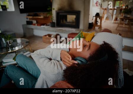 Vista ad alto angolo di una donna africana sorridente che si rilassa sul divano con le cuffie che ascoltano la musica con gli occhi chiusi Foto Stock