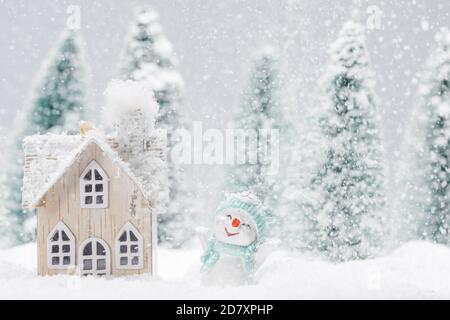 Piccolo pupazzo di neve decorativo vicino casa di legno in abeti foresta sotto caduta di neve Foto Stock