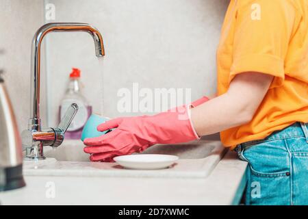 Una donna in abiti casual e guanti di gomma rosa sta lavando i piatti. Vista laterale. Lavori domestici. Foto Stock