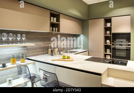 Cucina moderna ed elegante con bancone bar e sgabelli trasparenti moderni ed eleganti, elettrodomestici incorporati e armadietti beige neutri illuminati da soffitto incassato Foto Stock