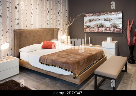 Interno di una elegante camera da letto moderna con ampio divano letto matrimoniale letto di stile con testiera marrone imbottita e tappeto con carta da parati marrone corrispondente o Foto Stock