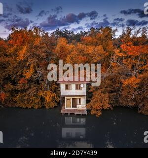 Tata, Ungheria - Vista aerea di una romantica cabina di pescatori sul lago Derito con cielo blu e fogliame autunnale al tramonto Foto Stock