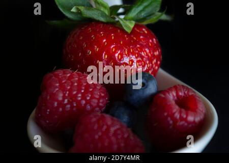 Bacche in un cucchiaio bianco.Fragola fresca, mirtillo e lampone.frutta fresca di foresta colorata su sfondo nero. Foto Stock