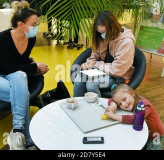 23 ottobre 2020, Sassonia-Anhalt, Köthen: Una madre si siede con i suoi due figli nell'ospedale pediatrico dell'ospedale universitario rilassato ad un tavolo nella caffetteria, su cui si trova un cercapersone (ricevitore cercapersone, in primo piano), che segnala la chiamata alla sala di consulenza con un ronzio. Al momento attuale, con un numero crescente di corona, il nuovo sistema cercapersone, con il quale i pazienti possono spostarsi fino a 500 metri di distanza, è destinato a equalizzare la situazione nelle aree di attesa. I pager dovrebbero permettere di mantenere le distanze anche in caso di una grande folla. La ricezione del cercapersone Foto Stock