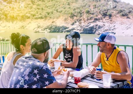 Gruppo di quattro amici con cappelli per proteggersi dal sole in una giornata di sole seduti intorno a un tavolo di legno mangiare, bere e divertirsi nel Foto Stock