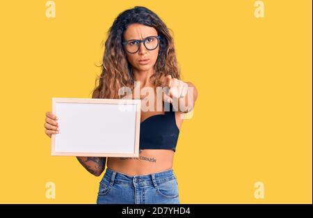 Giovane donna ispanica con tatuaggio che tiene vuota lavagna bianca che punta con il dito alla macchina fotografica e a voi, gesto sicuro che guarda serio Foto Stock