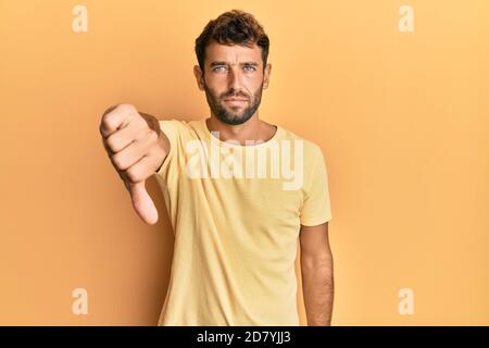 Bell'uomo con barba che indossa una camicia casual gialla sopra il giallo sfondo che guarda infelice e arrabbiato mostrando rifiuto e negativo con pollici verso il basso Foto Stock