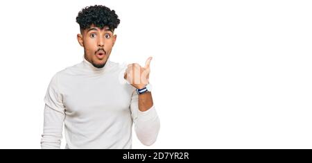 Giovane arabo che indossa un maglione invernale casual sorpreso puntando con il dito della mano a lato, bocca aperta stupito espressione. Foto Stock