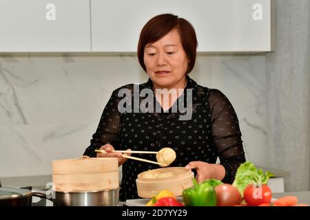 Donna asiatica matura che tiene un gnocco mentre si controlla un tradizionale vapore di bambù con verdure fuori fuoco. Cucina cinese tradizionale concetto. Foto Stock