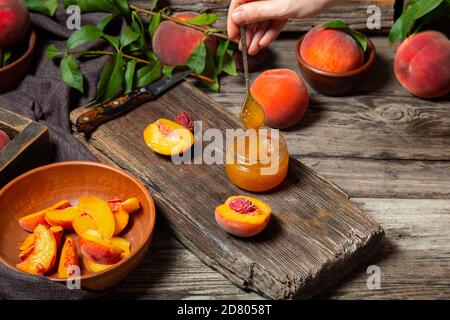 Il processo di preparazione della marmellata di pesche, cucinando il dessert di pesche su un tagliere rustico a mani femminili. Pesche frutti interi con foglie, pesche a metà Foto Stock