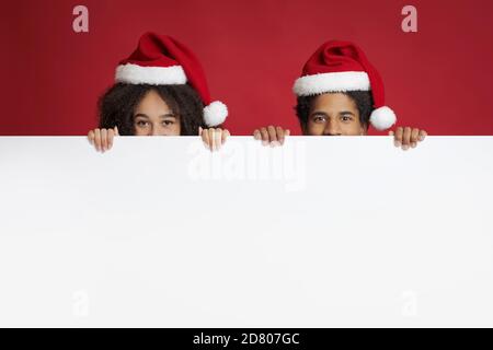 Offerte di Natale e Capodanno. Divertente marito afroamericano e moglie in cappelli peeking fuori da dietro poster Foto Stock