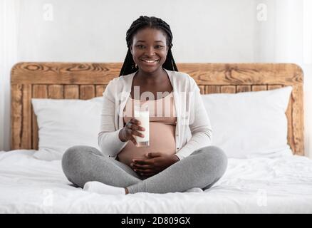 Donna africana in gravidanza felice bevendo latte mentre si siede sul letto A casa Foto Stock