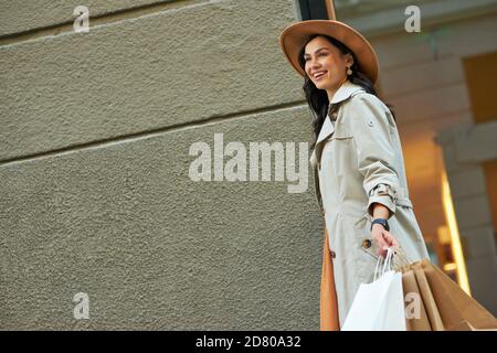 Visita boutique di moda. Giovane donna alla moda felice che indossa cappotto e cappello grigio autunno con borse per lo shopping passeggiando per le strade della città e sorridendo. Concetto di stile di vita delle persone Foto Stock